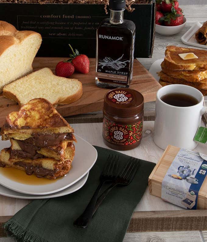 In the background, a loaf of brioche, with a bottle of syrup. In the foreground, a cup of tea, tea chest, a jar of chocolate hazelnut spread, and the finished product of brioche french toast,all on a wooden tray.