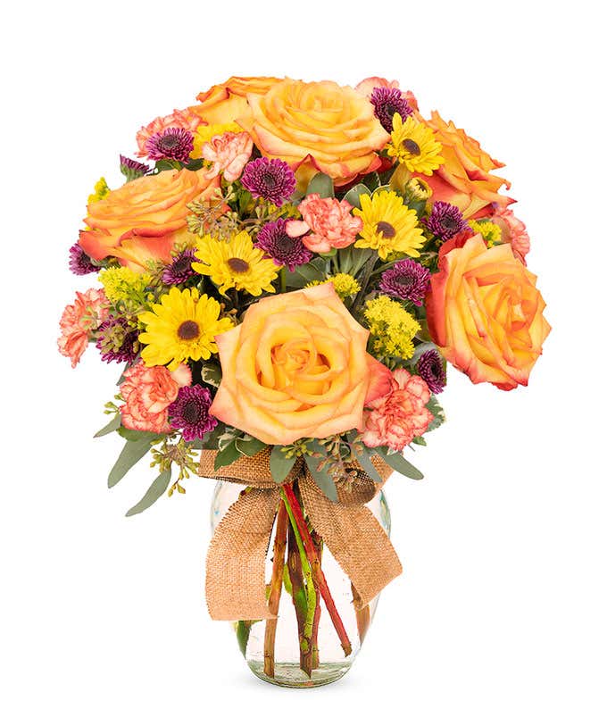 Festive fall bouquet full of orange roses, yellow daisies, and purple poms in a clear glass vase, wrapped in burlap ribbon.
