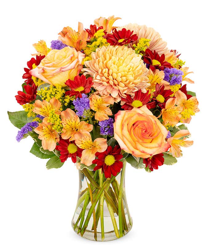 Roses, mums, and daisies in all the fall colors arranged into a tall clear vase.