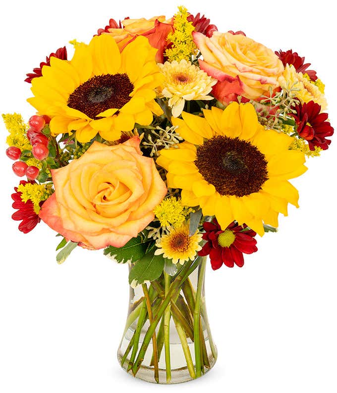 Sunflowers, roses, and daisies in shades of yellow, orange, and red, arranged in a clear glass vase.