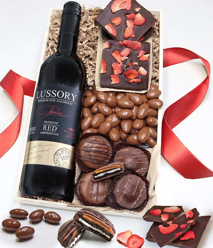 A gift box of chocolate covered Oreos topped with caramel, Dark Chocolate bark topped with dried strawberries, & a bag of chocolate covered almonds next to a bottle of non-alcoholic red wine