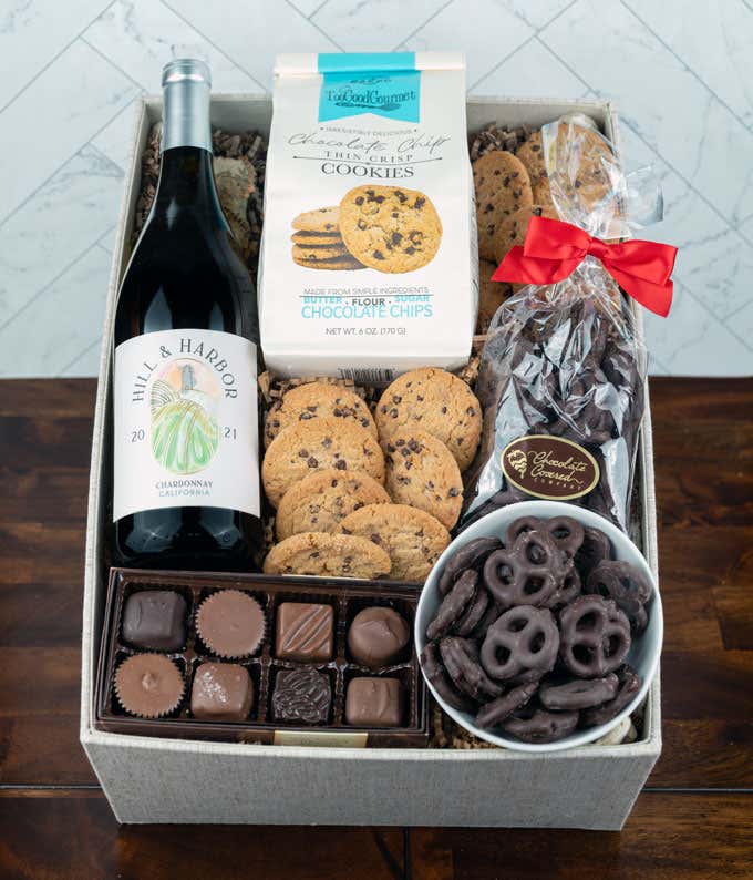 a bottle of white wine, two glasses of the wine, a bag of milk chocolate pretzel balls, a box of chocolate chip cookies, a box of assorted chocolates, and a cutting board all alid out on a white wooden table