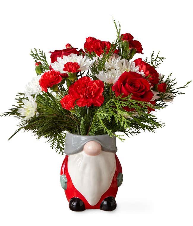 Red and white flowers arranged with assorted Christmas greens, placed into a festive gnome shaped ceramic vase