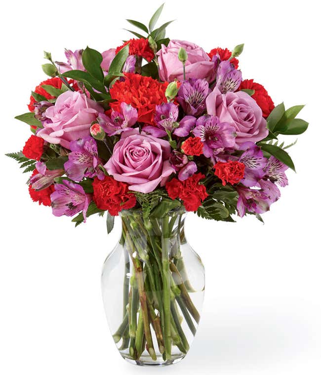 Lavender roses, purple alstroemeria, red carnations, and fresh floral greens in a clear glass vase