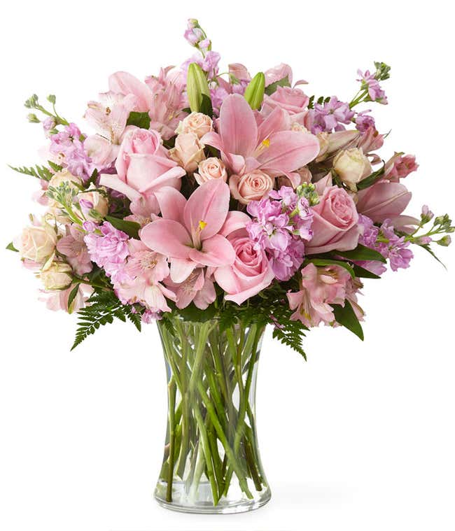 A bouquet of pink lilies, roses, stock and alstroemeria, with fresh floral greens in a tall cylinder vase