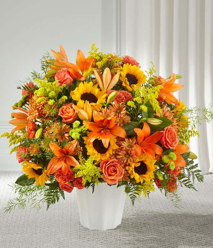 A standing basket of orange lilies, roses, and daisies, sunflowers, and fresh floral greens, in a white plastic urn