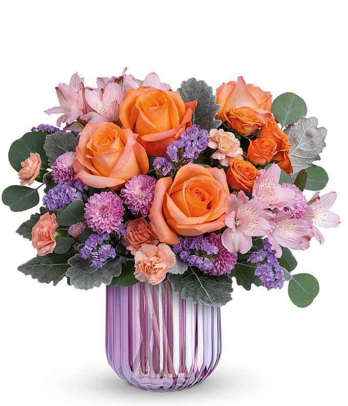 Short, round arrangement of orange roses and purple flowers, in a purple cylinder vase