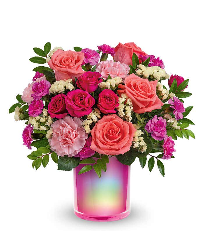 Short, round bouquet of coral, pink, and yellow flowers in a holographic cylinder vase