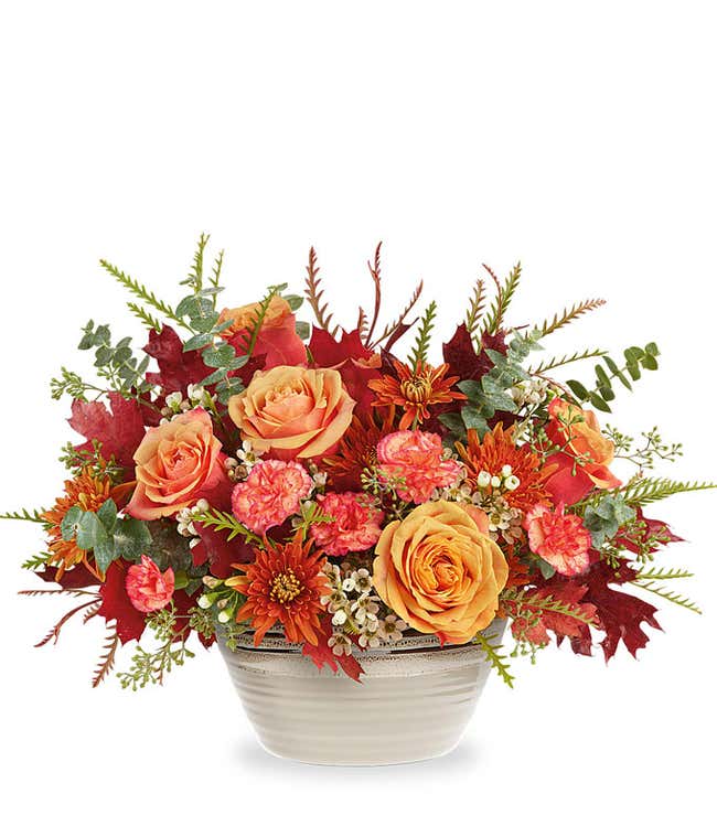 Light orange roses, orange carnations, bronze chrysanthemums, white waxflower, spiral &amp; seeded eucalyptus, grevillea, and preserved red oak leaves in a rustic beige bowl against a white background